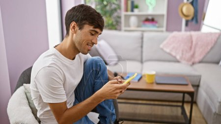 Téléchargez les photos : Jeune homme hispanique rayonnant assis à la maison, ayant un texto de souffle sur son smartphone, une image des temps plus heureux. son sourire éclatant crée une atmosphère joyeuse. profiter du confort intérieur. - en image libre de droit