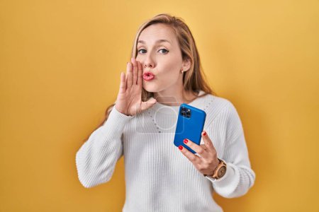 Photo for Young blonde woman using smartphone typing message hand on mouth telling secret rumor, whispering malicious talk conversation - Royalty Free Image