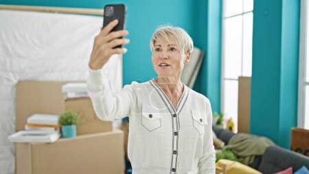 Photo for Middle age blonde woman having video call smiling at new home - Royalty Free Image