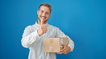 Photo for Young caucasian man holding package doing thumb up gesture over isolated blue background - Royalty Free Image