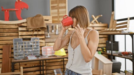 Photo for Serious young hispanic female carpenter sipping morning coffee, engaging in professional business call at bustling carpentry workshop - Royalty Free Image