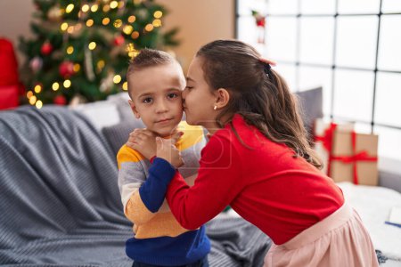Téléchargez les photos : Frère et sœur s'embrassent et s'embrassent debout près du sapin de Noël à la maison - en image libre de droit