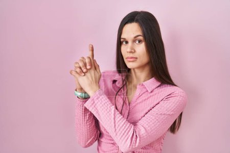 Téléchargez les photos : Young hispanic woman standing over pink background holding symbolic gun with hand gesture, playing killing shooting weapons, angry face - en image libre de droit