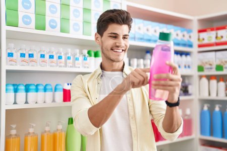 Téléchargez les photos : Jeune homme hispanique client souriant bouteille de shampooing de lecture confiant à la pharmacie - en image libre de droit