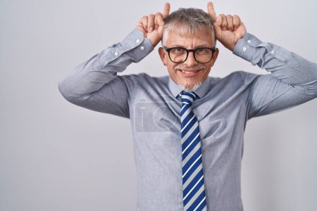 Téléchargez les photos : Homme d'affaires hispanique aux cheveux gris portant des lunettes faisant un geste drôle avec doigt au-dessus de la tête comme des cornes de taureau - en image libre de droit