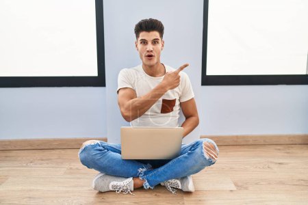 Foto de Joven hombre hispano usando portátil en casa sorprendido señalando con el dedo hacia un lado, boca abierta expresión asombrada. - Imagen libre de derechos