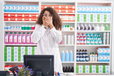 Téléchargez les photos : Femme hispanique avec les cheveux bouclés travaillant à la pharmacie pharmacie choqué couvrant la bouche avec les mains pour erreur. concept secret. - en image libre de droit