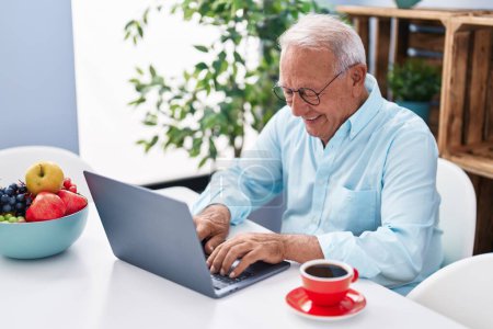 Photo for Senior grey-haired man using laptop and drinking coffee sitting on table at home - Royalty Free Image