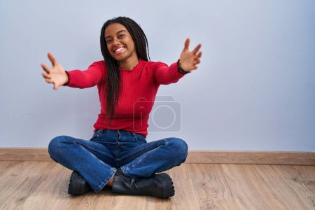 Téléchargez les photos : Jeune afro-américain avec des tresses assises sur le sol à la maison regardant la caméra souriant à bras ouverts pour un câlin. expression joyeuse embrassant le bonheur. - en image libre de droit