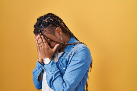 Téléchargez les photos : Femme afro-américaine avec des tresses debout sur fond jaune avec une expression triste couvrant le visage avec les mains tout en pleurant. concept de dépression. - en image libre de droit