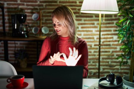 Téléchargez les photos : Femme blonde utilisant un ordinateur portable la nuit à la maison expression dégoûtée, mécontente et effrayée faisant visage dégoûtant parce que réaction d'aversion. les mains levées - en image libre de droit