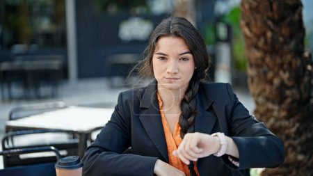 Téléchargez les photos : Jeune belle hispanique femme ouvrière regardant horloge avec une expression sérieuse à la terrasse du café - en image libre de droit
