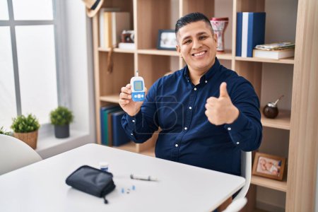 Téléchargez les photos : Jeune homme hispanique tenant dispositif glucomètre souriant heureux et positif, pouce levé faire excellent et signe d'approbation - en image libre de droit