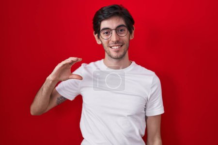 Téléchargez les photos : Jeune homme hispanique debout sur fond rouge geste souriant et confiant avec la main faisant signe de petite taille avec les doigts regardant et la caméra. concept de mesure. - en image libre de droit