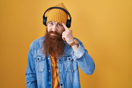 Foto de Hombre caucásico con barba larga escuchando música usando audífonos que apuntan infelices a la espinilla en la frente, fea infección de la cabeza negra. acné y problemas de piel - Imagen libre de derechos