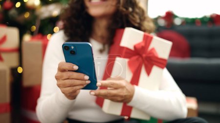 Foto de Mujer hispana de mediana edad usando teléfono inteligente sentado en el suelo por árbol de Navidad en casa - Imagen libre de derechos