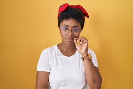 Téléchargez les photos : Jeune femme afro-américaine debout sur fond jaune bouche et lèvres fermées comme zip avec les doigts. secret et silencieux, tabou parlant - en image libre de droit