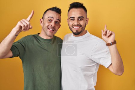 Téléchargez les photos : Couple homosexuel debout sur fond jaune souriant étonné et surpris et pointant vers le haut avec les doigts et les bras levés. - en image libre de droit