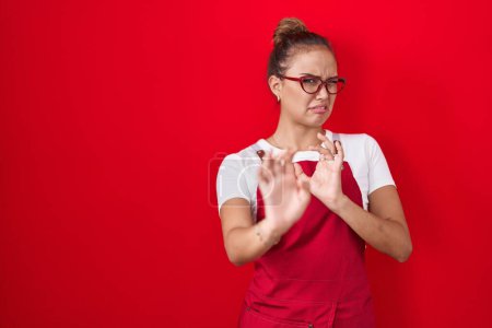Photo for Young hispanic woman wearing waitress apron over red background disgusted expression, displeased and fearful doing disgust face because aversion reaction. with hands raised - Royalty Free Image