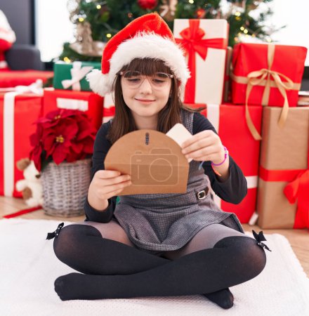 Foto de Adorable chica hispana sosteniendo Santa Claus carta sentada en el suelo por el árbol de Navidad en casa - Imagen libre de derechos