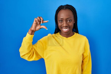 Téléchargez les photos : Belle femme noire debout sur fond bleu geste souriant et confiant avec la main faisant signe de petite taille avec les doigts regardant et la caméra. concept de mesure. - en image libre de droit