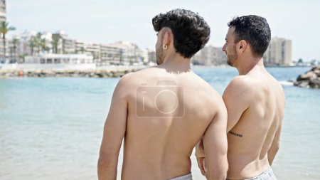 Téléchargez les photos : Deux hommes couple touristique debout vers l'arrière avec les mains ensemble à la plage - en image libre de droit
