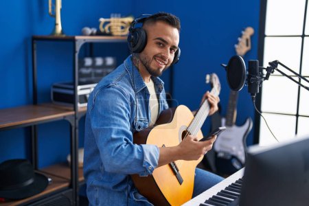 Photo for Young hispanic man artist using smartphone playing classical guitar at music studio - Royalty Free Image