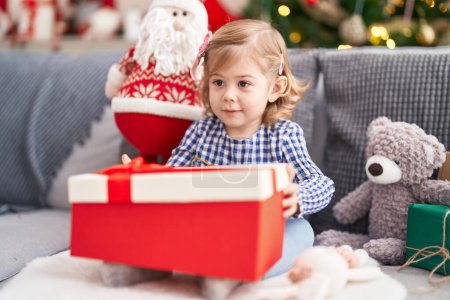 Photo for Adorable hispanic girl holding gift sitting on sofa by christmas tree at home - Royalty Free Image