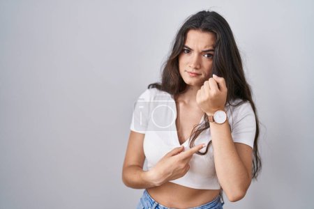 Photo for Young teenager girl standing over white background in hurry pointing to watch time, impatience, looking at the camera with relaxed expression - Royalty Free Image