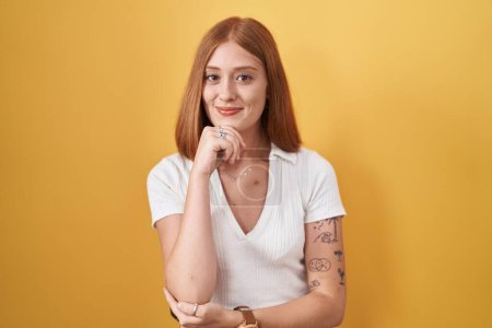 Photo for Young redhead woman standing over yellow background looking confident at the camera smiling with crossed arms and hand raised on chin. thinking positive. - Royalty Free Image