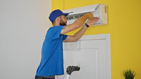 Téléchargez les photos : Jeune homme hispanique technicien réparer la machine de climatisation debout sur l'échelle à la maison - en image libre de droit