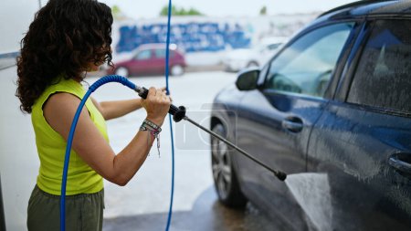 Téléchargez les photos : Moyen Age femme hispanique lavage de voiture avec lave-pression à la station de lavage de voiture - en image libre de droit