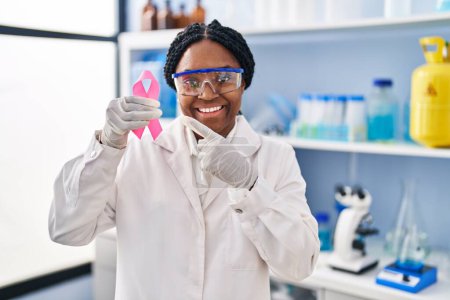 Photo for African american woman working at scientist laboratory holding pink ribbon smiling happy pointing with hand and finger - Royalty Free Image
