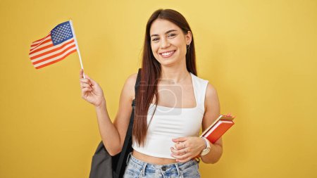 Téléchargez les photos : Jeune belle étudiante hispanique souriante confiante tenant des livres et drapeau des États-Unis sur fond jaune isolé - en image libre de droit