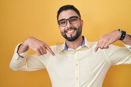 Téléchargez les photos : Jeune homme hispanique portant des vêtements de travail et des lunettes regardant confiant avec le sourire sur le visage, se montrant du doigt fier et heureux. - en image libre de droit