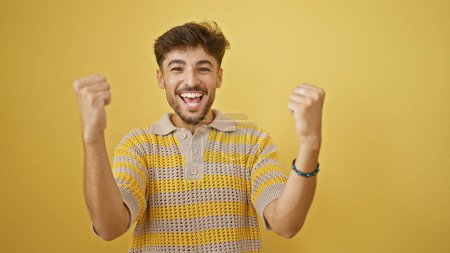 Téléchargez les photos : Jeune homme arabe confiant savourant un pur bonheur, célébrant joyeusement sa victoire avec un sourire éclatant, debout sur un fond jaune isolé vibrant. - en image libre de droit