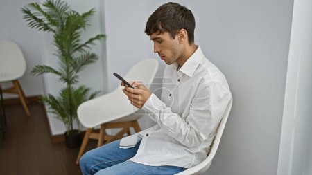Téléchargez les photos : Sérieux jeune hispanique, un professionnel à succès, assis détendu sur une chaise, absorbé par les textos sur son smartphone, attendant une réunion d'affaires importante dans une salle d'attente spacieuse - en image libre de droit