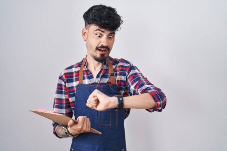 Téléchargez les photos : Jeune homme hispanique avec barbe portant tablier de serveur tenant presse-papiers regardant l'heure de la montre inquiet, peur de se retrouver en retard - en image libre de droit