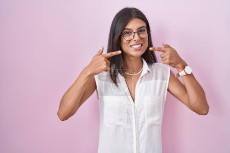 Téléchargez les photos : Brunette jeune femme debout sur fond rose portant des lunettes souriant gai montrant et pointant avec les doigts dents et la bouche. concept de santé dentaire. - en image libre de droit