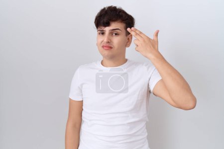 Téléchargez les photos : Jeune homme non binaire portant un t-shirt blanc décontracté tirant et se suicidant pointant la main et les doigts vers la tête comme une arme à feu, geste de suicide. - en image libre de droit