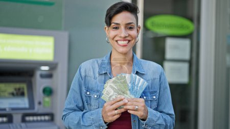 Foto de Joven hermosa mujer hispana sonriendo confiada sosteniendo billetes chilenos en cajero de banco - Imagen libre de derechos