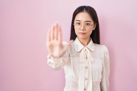 Foto de Joven mujer china de pie sobre fondo rosa haciendo dejar de cantar con la palma de la mano. expresión de advertencia con gesto negativo y grave en la cara. - Imagen libre de derechos