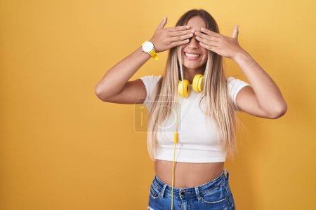 Téléchargez les photos : Jeune femme blonde debout sur fond jaune portant des écouteurs couvrant les yeux avec les mains souriantes gaies et drôles. concept aveugle. - en image libre de droit