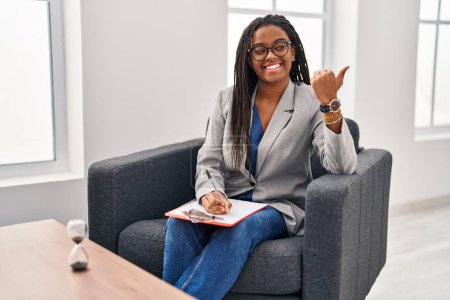 Téléchargez les photos : Jeune afro-américain avec des tresses travaillant au bureau de consultation souriant avec le visage heureux regardant et pointant vers le côté avec le pouce vers le haut. - en image libre de droit