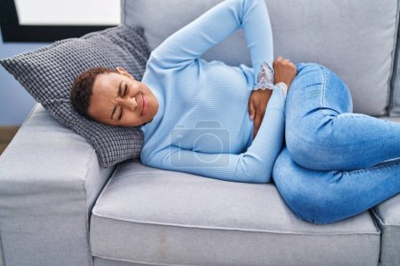 Foto de Mujer afroamericana que sufre de dolor menstrual tumbada en un sofá en casa - Imagen libre de derechos