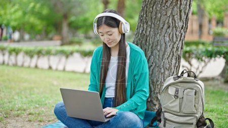 Téléchargez les photos : Jeune femme chinoise utilisant ordinateur portable et écouteurs assis sur le sol au parc - en image libre de droit