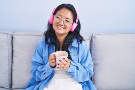 Foto de Joven mujer china escuchando música tomando café en casa - Imagen libre de derechos