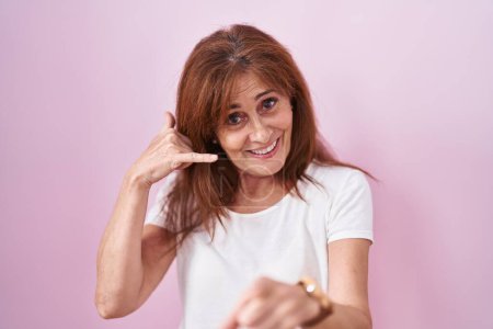 Téléchargez les photos : Femme d'âge moyen debout sur fond rose souriant faire parler au téléphone geste et vous pointant du doigt. Appelle-moi. - en image libre de droit