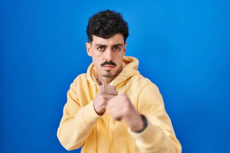 Photo for Hispanic man standing over blue background ready to fight with fist defense gesture, angry and upset face, afraid of problem - Royalty Free Image
