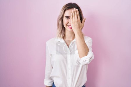Téléchargez les photos : Young beautiful woman standing over pink background covering one eye with hand, confident smile on face and surprise emotion. - en image libre de droit